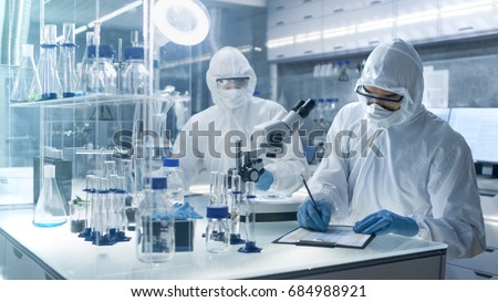 In a Secure High Level Laboratory Scientists in a Coverall Conducting a Research. Chemist Writes Down Experiment Results.