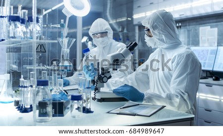 In a Secure High Level Laboratory Scientists in a Coverall Conducting a Research. Chemist Adjusts Samples and Discuss Experiment with Collegue.