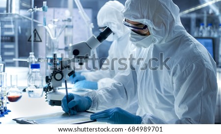 In a Secure High Level Laboratory Scientists in a Coverall Conducting a Research. Chemist with His Colleague Writes Down Experiment Results.