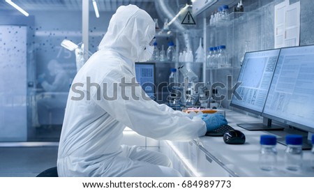 In a Secure High Level Laboratory Scientists in a Coverall Conducting a Research. Biologist Analyzes Results on a Computer.