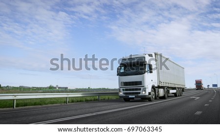 Speeding White Semi Truck with Cargo Trailer Drives on the Highway. Truck is First in the Column of Heavy Vehicles, Sun is Shining.