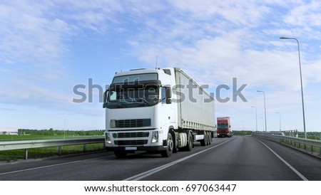 Speeding White Semi Truck with Cargo Trailer Drives on the Highway. Truck is First in the Column of Heavy Vehicles, Sun is Shining.
