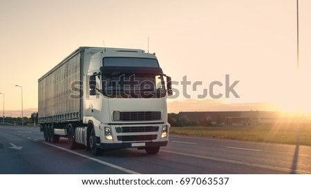 Front-View of Semi-Truck with Cargo Trailer Driving on a Highway. He\'s Speeding Through Industrial Warehouse Area with Sunset in the Background.