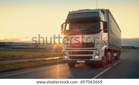 Front-View of Semi-Truck with Cargo Trailer Driving on a Highway. He\'s Speeding Through Industrial Warehouse Area with Sunset in the Background.