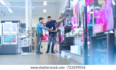 In the Electronics Store Professional Consultant Shows Latest TV\'s to a Young Man, They Talk about Specifications and What Model is Best for Young Man\'s Home. Store is Bright and Has Latest Models.