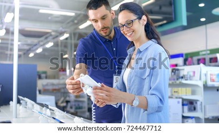 In the Electronics Store Consultant Gives Professional Advice to a Young Woman, She Considers Buying New Tablet Computer and Needs Expert Opinion. Store is Modern, Bright and Has all the New Devices.