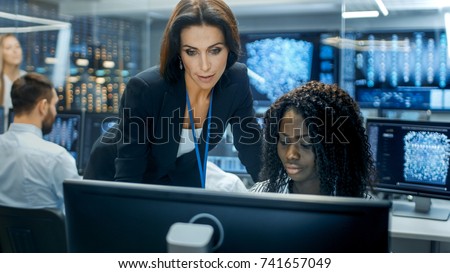 Female Team Leader Consults Young Computer Engineer. They Work in a Crowded Office on a Neural Network/ Artificial Intelligence Project.