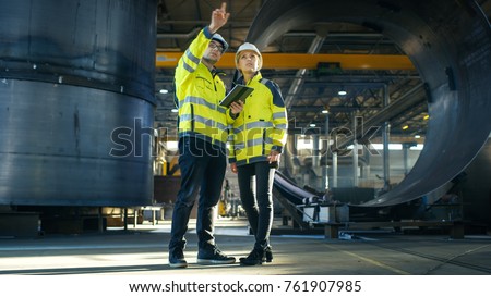 Male and Female Industrial Engineers in Hard Hats Discuss New Project while Using Tablet Computer. They're Making Calculated Engineering Decisions.They Work at the Heavy Industry Manufacturing Factory