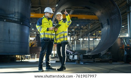 Male and Female Industrial Engineers in Hard Hats Discuss New Project while Using Tablet Computer. They\'re Making Calculated Engineering Decisions.They Work at the Heavy Industry Factory.