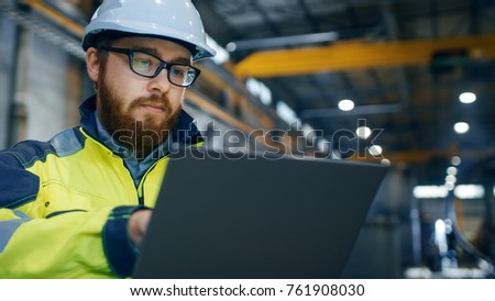 Industrial Engineer in Hard Hat Wearing Safety Jacket Uses Touchscreen Laptop. He Works at the Heavy Industry Manufacturing Factory. In the Background Welding/ Metalworking Processes are in Progress.