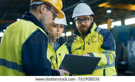 Three Industrial Engineers Talk with Factory Worker while Using Laptop. They Work at the Heavy Industry Manufacturing Facility.