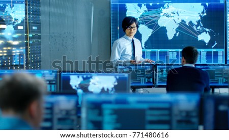 In the System Control Room Manager Holds a Briefing for His Staff Members. They\'re Work in Data Center and are Surrounded by Multiple Screens Showing Maps, Logistics Data.