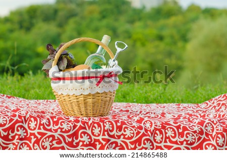 Decorated picnic basket with a bottle of white wine, corkscrew, buns and bunch of basil on red tablecloth, green landscape