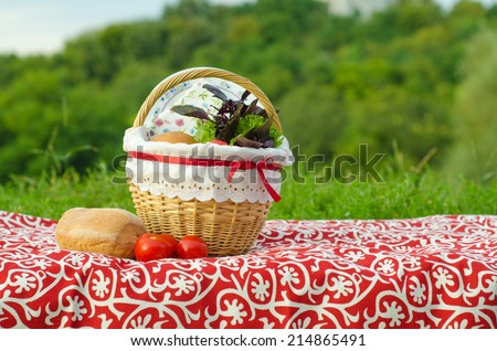 Decorated picnic basket with plate, buns and bunch of basil and salad, tomatoes on red tablecloth, green landscape