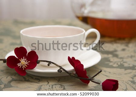 A cup of tea in a white china teacup accented with a red Japanese flower blossoms with a glass tea pot in the background.
