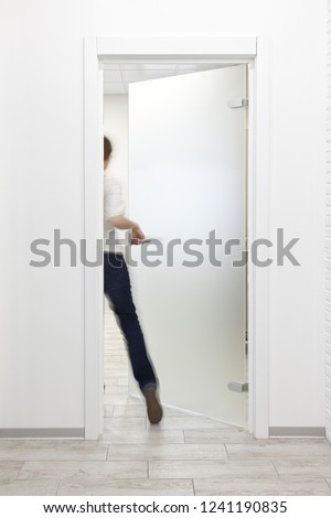 Young woman entering a room in modern office with minimalist white interior through frosted glass door. Woman is motion blurred