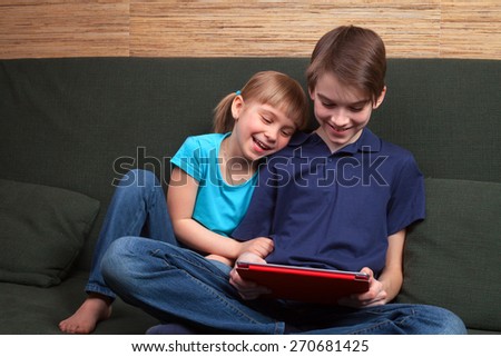 Children wearing casual clothes playing or watching a movie on a touch pad at home sitting on a green sofa. Boy and girl are half-siblings. Brother is holding tablet with red cover.