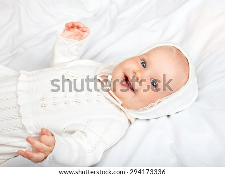 Close-up shot of six month baby girl wearing white christening clothes laying on a bed smiling