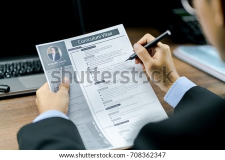 Businessman using the pen to reviewing or checking the Curriculum Vitae or CV of the candidate before interviewing with a laptop and phone on the wood table in background