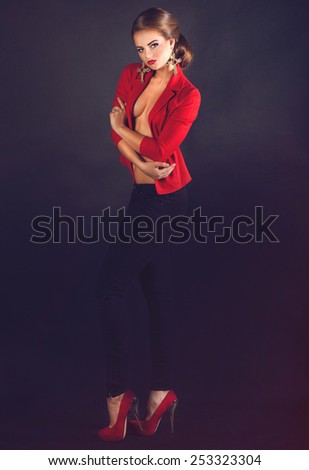 Beautiful woman posing full length with naked breasts in red jacket and black pants with crosses earrings on black background with copyspace