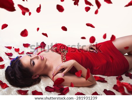 Sexy beautiful brunette girl in red dress laying on a bed under romantic falling rose petals