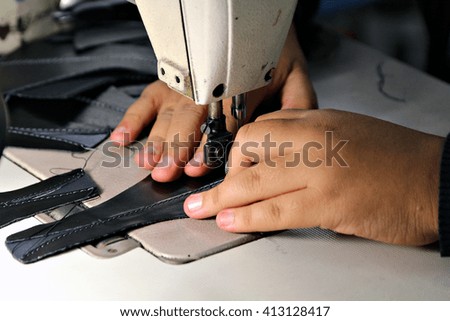 Closeup of a woman\'s hands as she operates an industrial sewing machine in a shoe factory, hand-stitching a the leather upper of a shoe. Shallow depth of field.