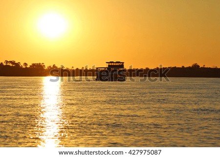 A houseboat cruises the Zambezi River, the border between Zimbabwe and Zambia, Africa, at sunset as the tourists on board enjoy sundowner cocktails. Sunset Cruise. Focus on boat.