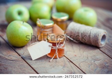 Apple jam in a small glass jar and green apples on wooden table. Blank label provides copy space for a message