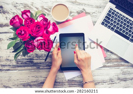 Bouquet of roses, cup of coffee, female hands with tablet and laptop on old wooden table. Top view. Toned image