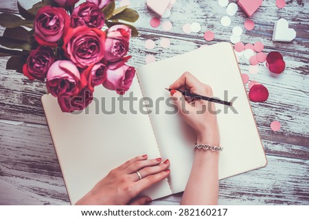 Female hands writing in open notebook and bouquet of roses on old wooden table. Top view. Toned image