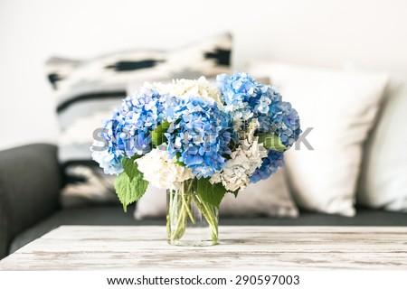 Bouquet of hortensia flowers on modern wooden coffee table and cozy sofa with pillows. Living room interior and home decor concept
