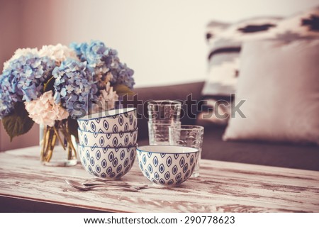Bouquet of hortensia flowers and glass bowls on modern wooden coffee table and cozy sofa with pillows. Living room interior and home decor concept. Toned image