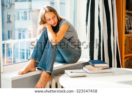 A sad teenage girl sits on window sill instead of reading books on the table