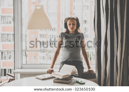 A sad teenage girl sits on window sill listening to music instead of reading books on the table. Toned image