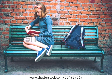 A teenage girl is reading on a bench with brick wall in the background. Toned image