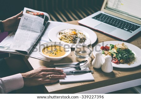 Business woman sits in cafe and reads the newspaper. Three plates with lunch dishes on wooden table. Caesar salad, pumpkin soup and pasta carbonara. Toned image