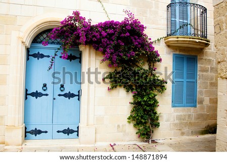 Old street in Malta with purple flowers crawling up a wall near a blue door.