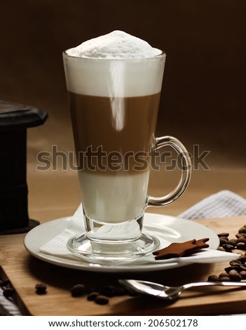 Latte macchiato with a white napkin, white plate, spoon on a wooden board, decorated with coffee beans, cinnamon stars and vintage coffee grinder and Turkish coffee pot in the back.