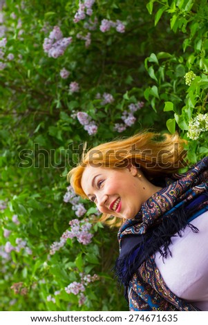 Young woman in motion on the background of blooming lilacs, sparkling emotions, flirting, joy, laughter, life. The girl pavlopasadsky Russian scarf and holding a fan. Spring.