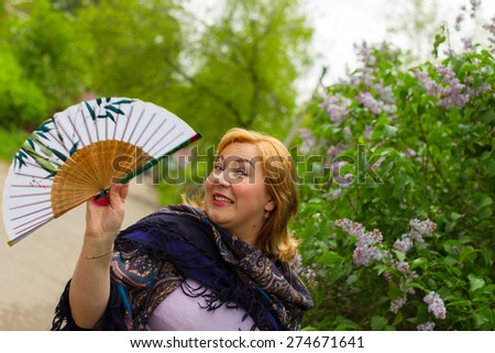 Young woman in motion on the background of blooming lilacs, sparkling emotions, flirting, joy, laughter, life. The girl pavlopasadsky Russian scarf and holding a fan. Spring.