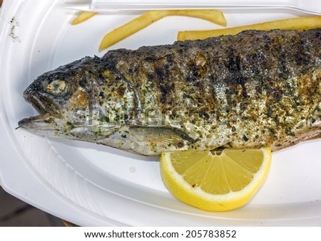 grilled trout with a slice of lemon and some french fries on a white plastic plate in an open door restaurant