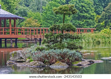 Fragment of a japanese garden with an island, artificially shaped trees and red bridge with viewing pavilion in background