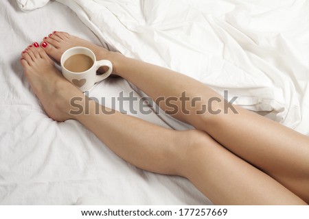 A cup of coffee or hot chocolate and female feet on a white sheets.