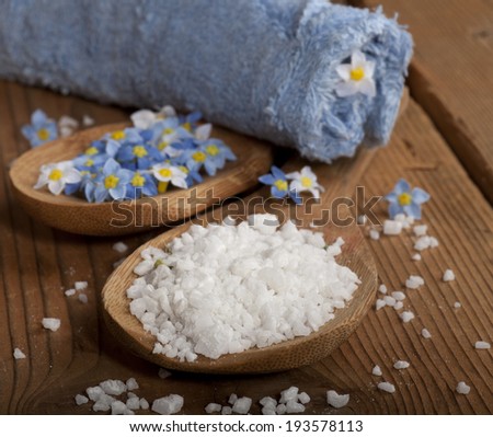 Spa background with sea salt in wooden spoon, flowers and rolled towel on wooden background.