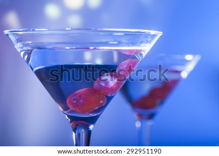 Two glasses of martini cocktail with red cherries on a mixed color background.