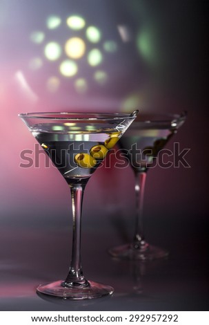 Two glasses of martini cocktail with green olives on a mixed color background.