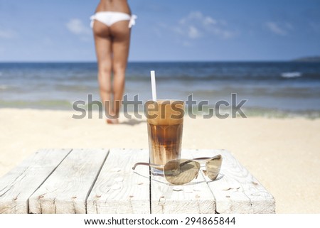 Iced coffee and glasses on a wooden table on the beach. In the distance woman in swinsuit and the sea.
