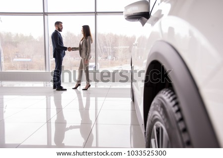Wide angle portrait of car salesman shaking hands with woman buying new car in dealership showroom, car wheel in foreground, copy space