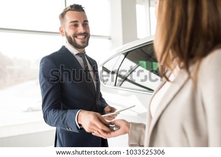 Portrait of handsome car salesman giving car keys to young woman standing next to white shiny luxury car in dealership showroom