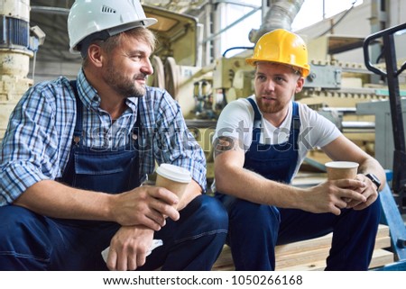 Portrait of two workers wearing hardhats taking break from work drinking coffee and resting sitting on pack of wood in workshop